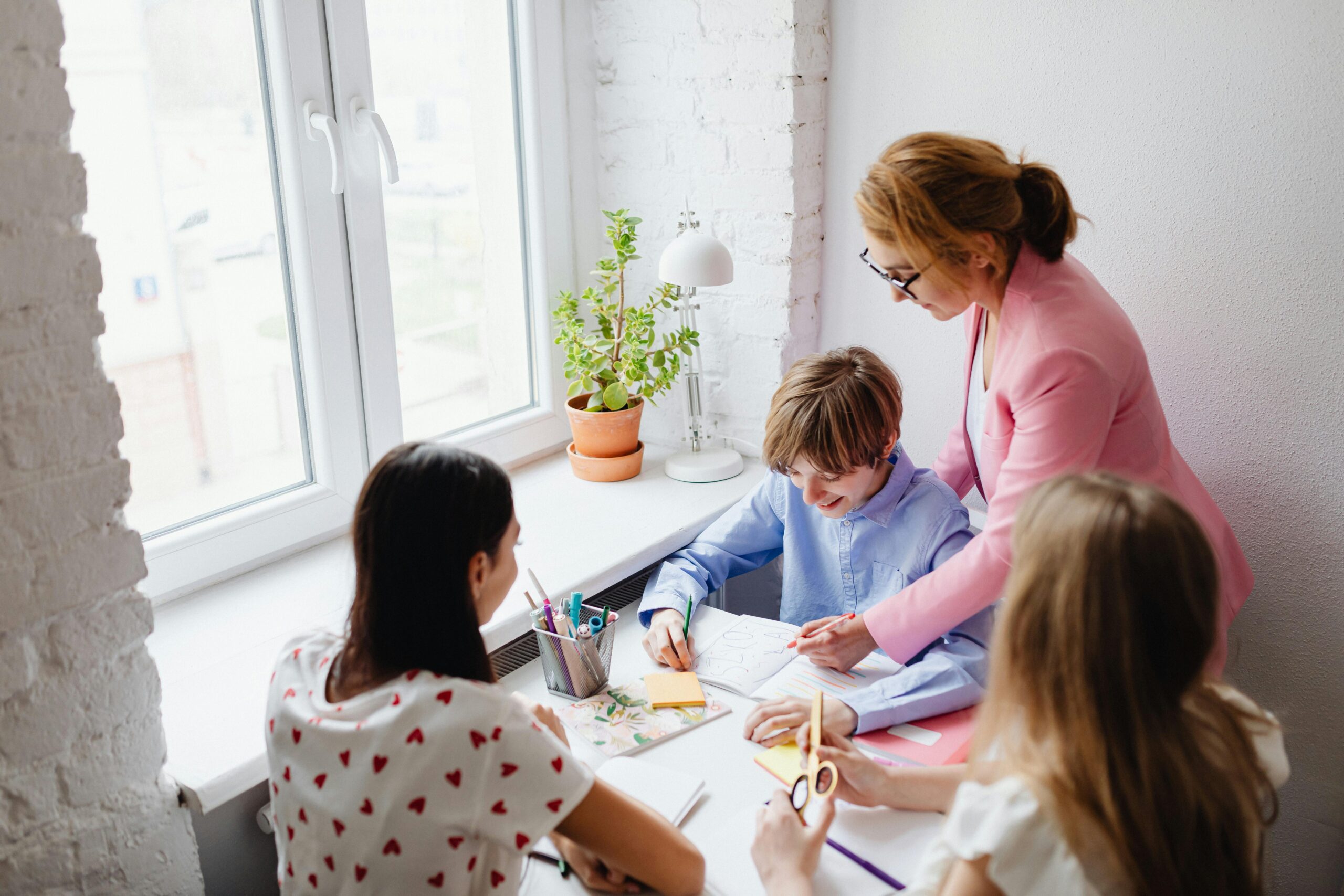 A teacher assists students with schoolwork around a desk in a bright classroom.