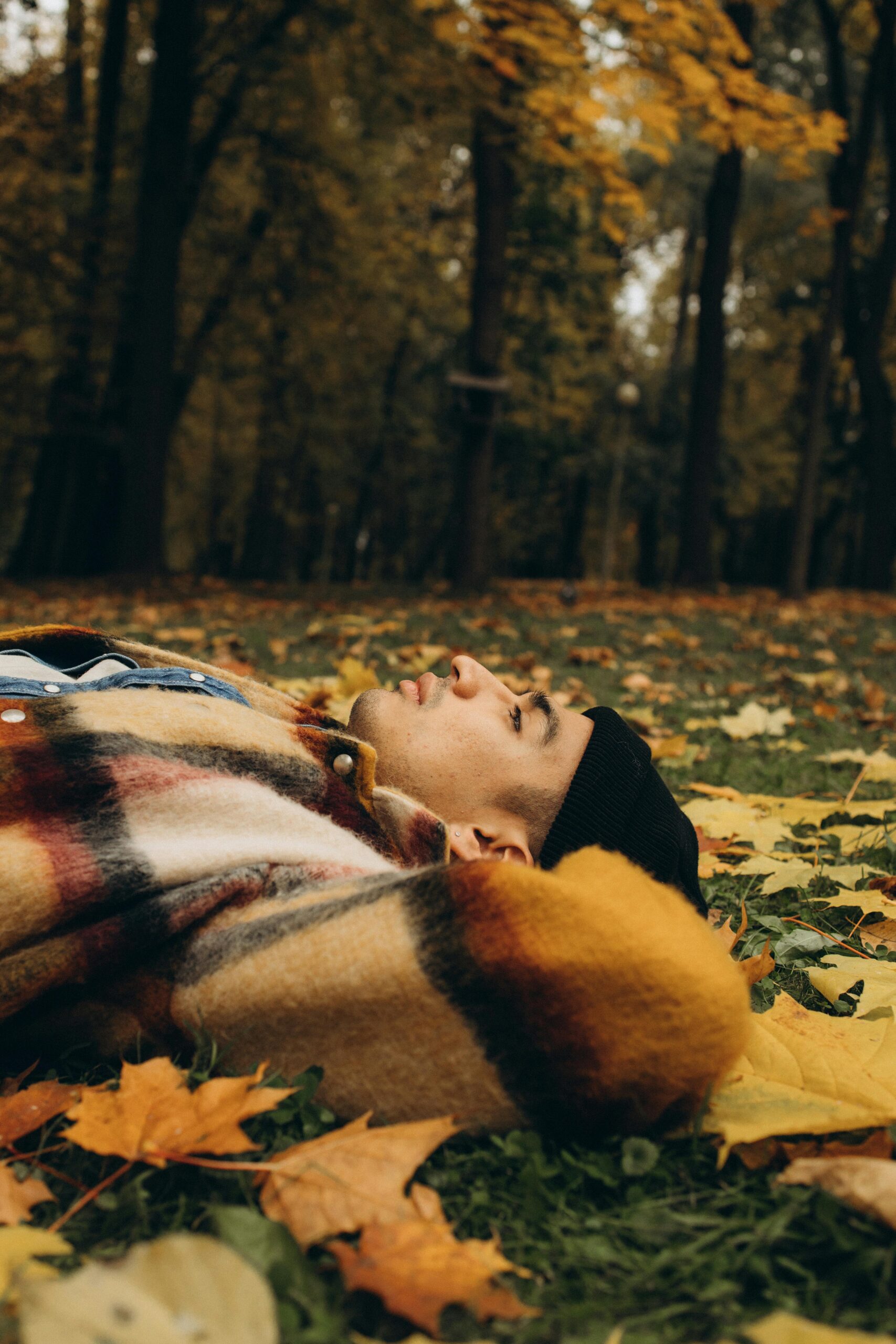 A young man in a plaid jacket rests on autumn leaves in a serene forest setting.